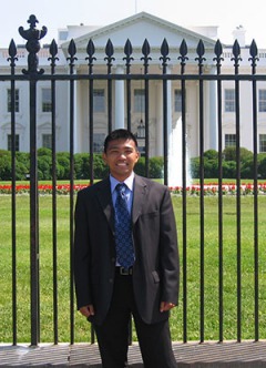 Dark skinned man, dark hair, dark suit, blue patterned tie, standing in front of fence, White House in background.