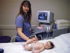 Light skinned woman, dark hair, blue t-shirt, standing in front of ultrasound screen, scoping baby (test doll) on bed.