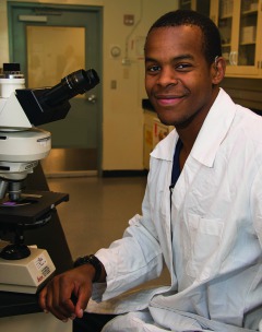 Dark skinned young man wearing lab coat sitting in front of telescope.