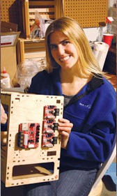Light skinned woman, long blond hair, dark sweater, holding circuit board.