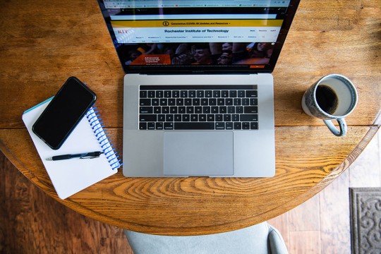 Overhead view of open laptop on kitchen table next to mug of coffee, notepad and cell phone.