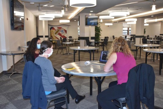three employees wearing face masks sitting at a round table watching a presentation on a laptop.