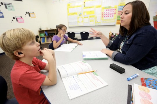 elementary student with a cochlear implant sitting across from a teacher at a table with a math workbook.
