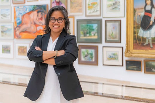 art gallery director standing with her arms crossed in front of a wall of paintings.