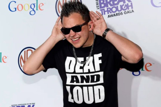 Deaf rapper Sean Forbes wearing sunglasses and a T shirt that says Deaf and Loud. He is holding his hands behind his ears.
