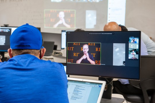 a college student facing a computer screen that shows a video call with a person using sign language.