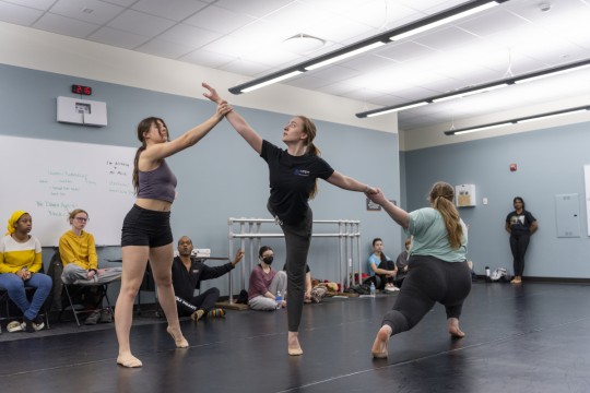 Image of students in a dance studio with some practicing and others sitting behind them watching. 