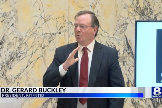 "Dr. Gerard Buckley, president of RIT/NTID, speaks using American Sign Language in front of a marble wall, with a screen displaying content beside him.