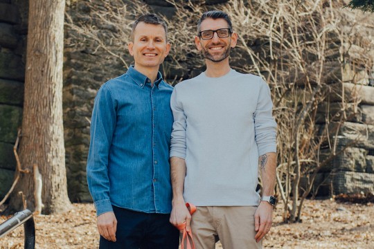 Two smiling men pose outdoors in front of a stone wall and leafless branches. One wears a denim shirt, while the other wears a white sweater and holds a red leash.