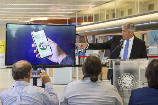 A man in a suit gestures to a screen showing an image of the AIRA A S L smartphone app during a news conference.