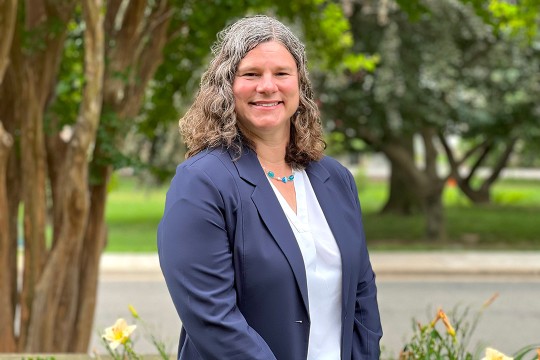 a woman with curly gray hair stands in a park like setting in a navy blue suit.