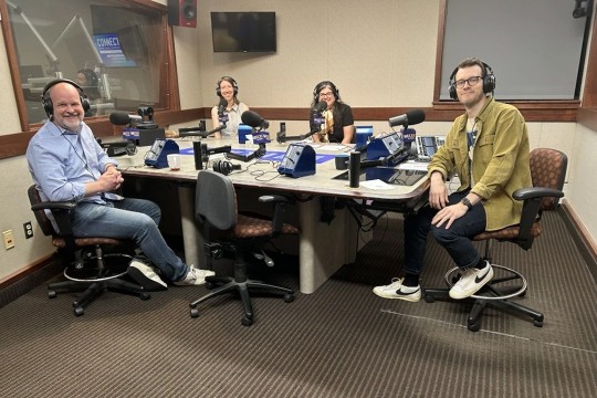 4 people sit around a table with headphones in front of microphones.