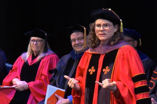 a woman in glasses and curly brown hair stands in red regalia for the university on a stage.