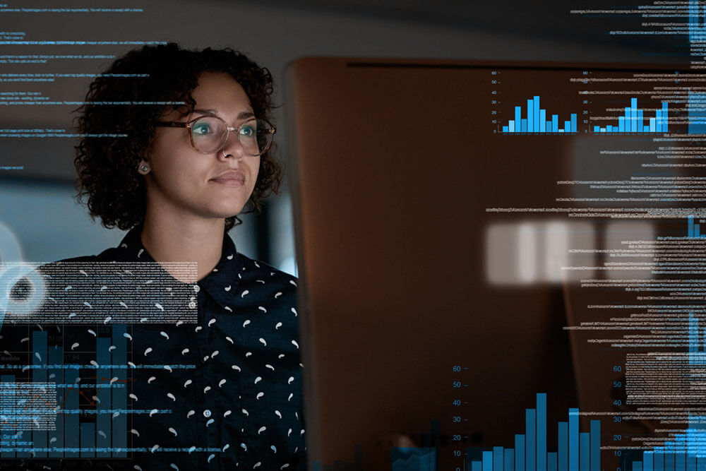 A woman with glasses sits at a desktop monitor with images of charts and text overlayed over the photo.
