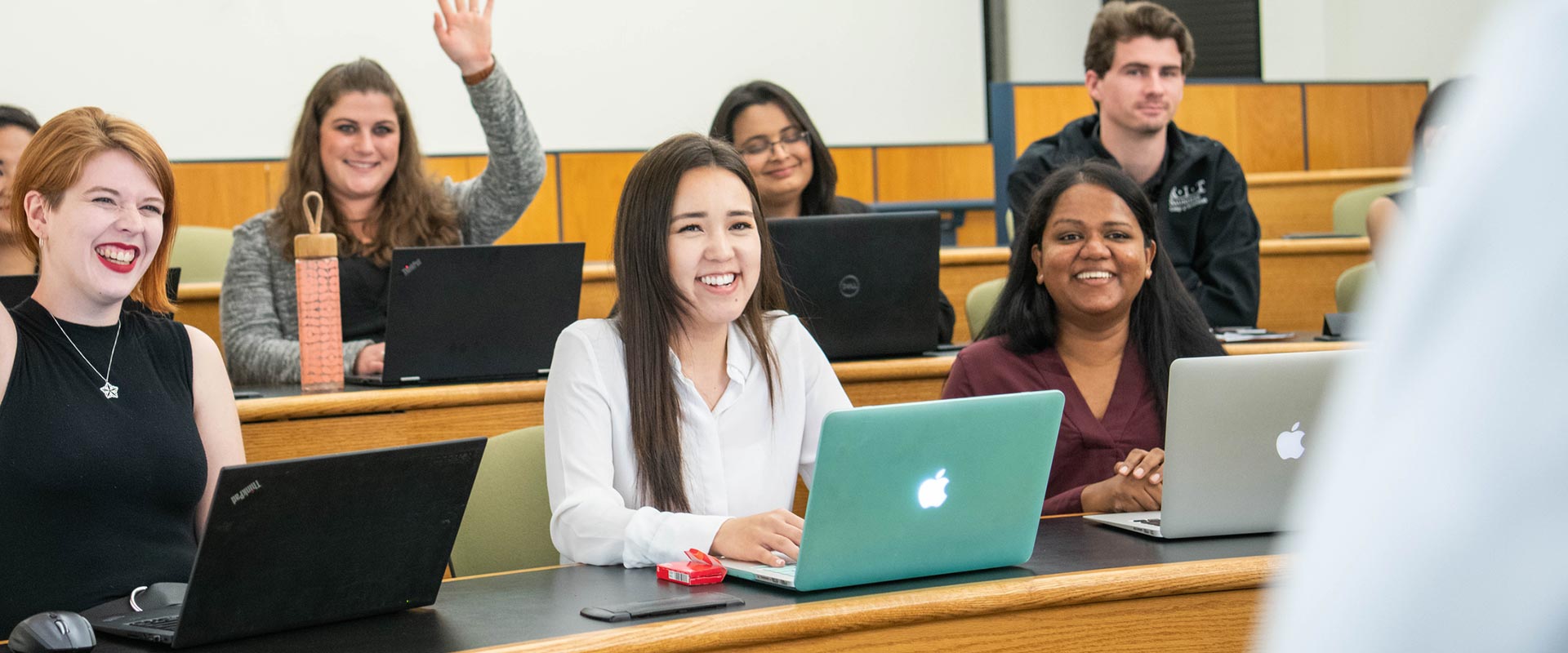 students smiling and sitting in a lecture hall, with one student raising her hand.