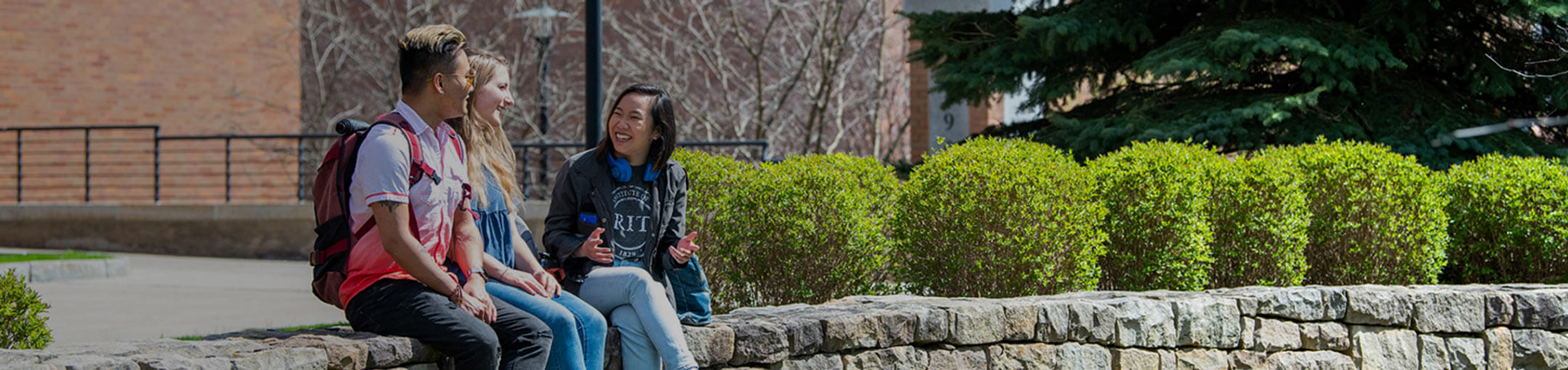 Three RIT students sitting on a stone wall in a quad on campus.