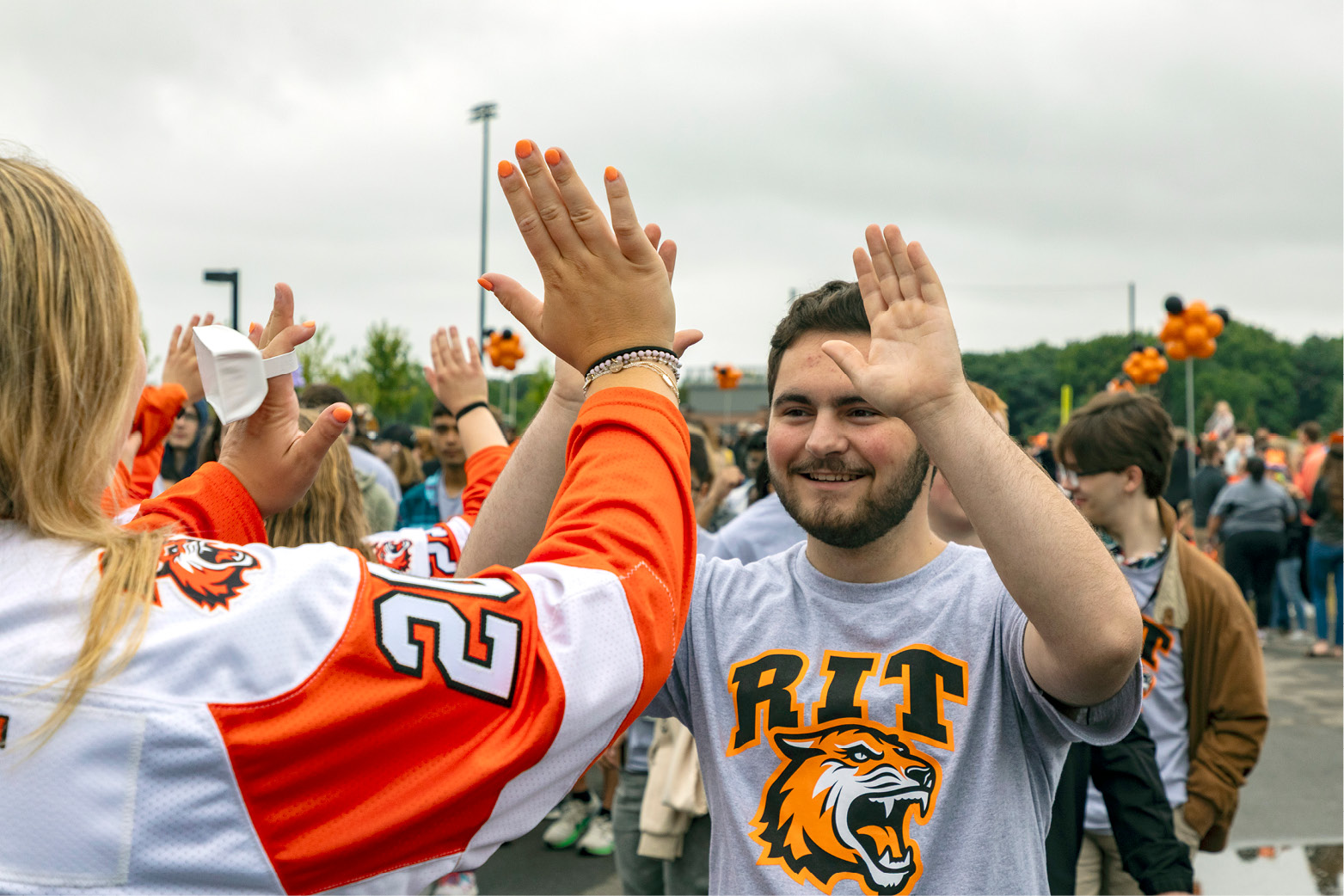 Two students exchange a high-five at a crowded outdoor campus event, with other attendees, orange decorations, and RIT apparel visible in the background.
