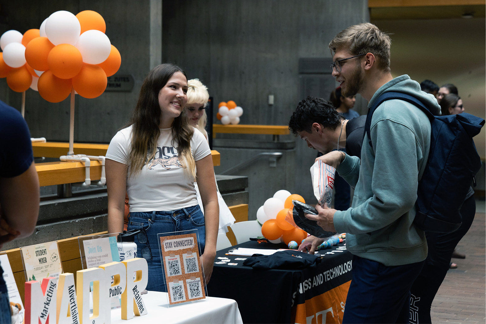 A student stands behind an information table at an indoor event, talking with another student while promotional materials and orange-and-white balloon decorations are displayed.