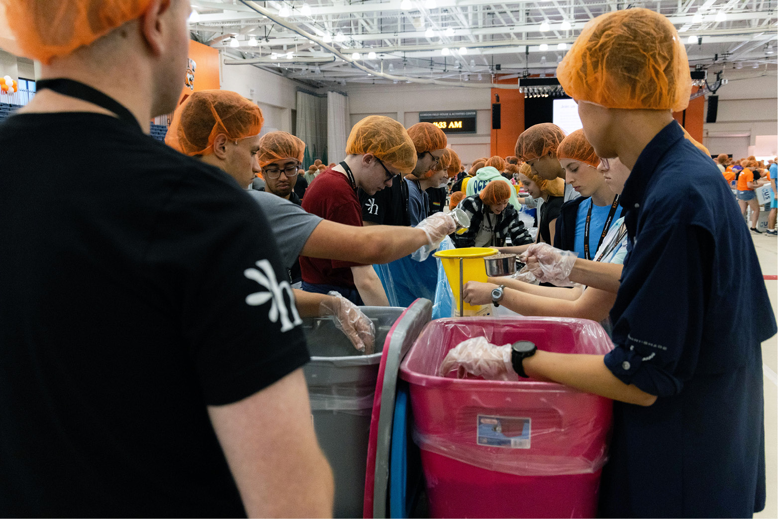 Students wearing hair nets and gloves stand around tables indoors, assembling food items into large bins as part of a volunteer or service activity.