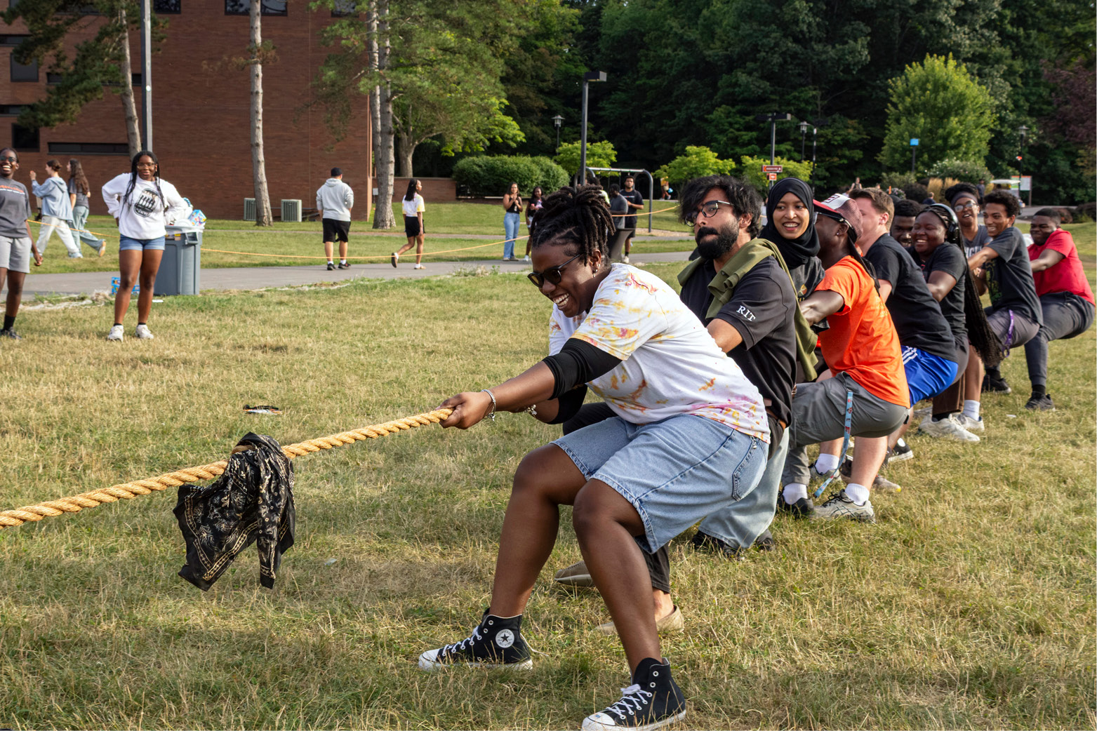 A group of students participate in a tug-of-war on a grassy campus lawn, pulling a thick rope during an outdoor activity with buildings and onlookers in the background.