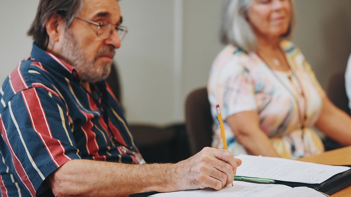 Older man in a striped shirt taking notes with a pencil during a classroom session.