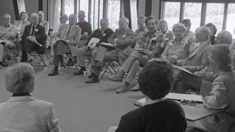 black and white photo of older adults seated in chairs in a circle listening to a speaker.