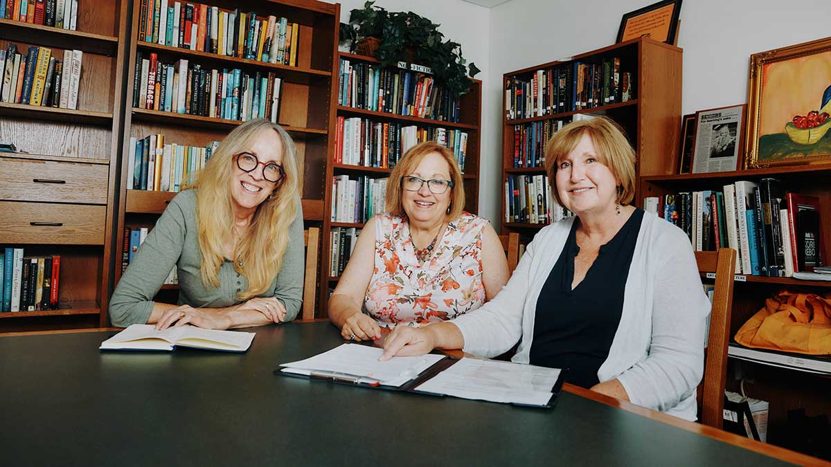 Three women seated at a table in a library, smiling and looking at a binder.