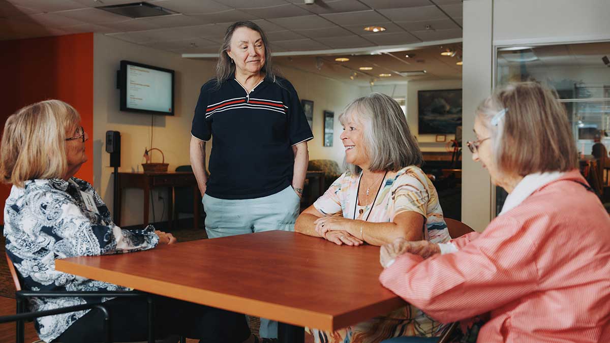Three seated women and one standing woman talking at a square table inside a lounge area.