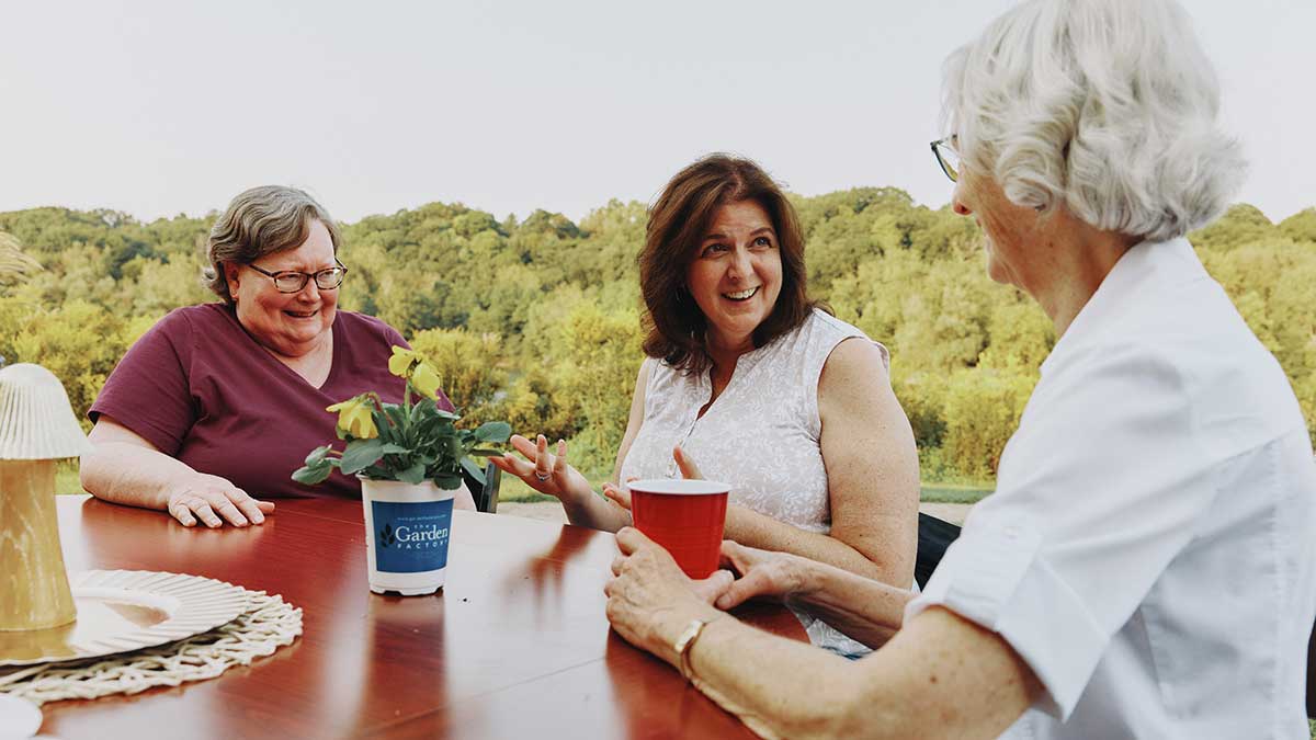 Three women sitting outdoors at a table, smiling and talking during a social gathering.