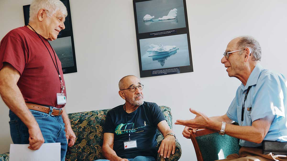 Three older men having a conversation in a room with framed iceberg photos on the wall.