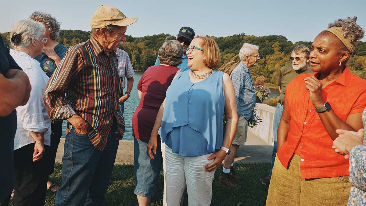 Group of older adults gathered outdoors near water, smiling and talking during a social event.
