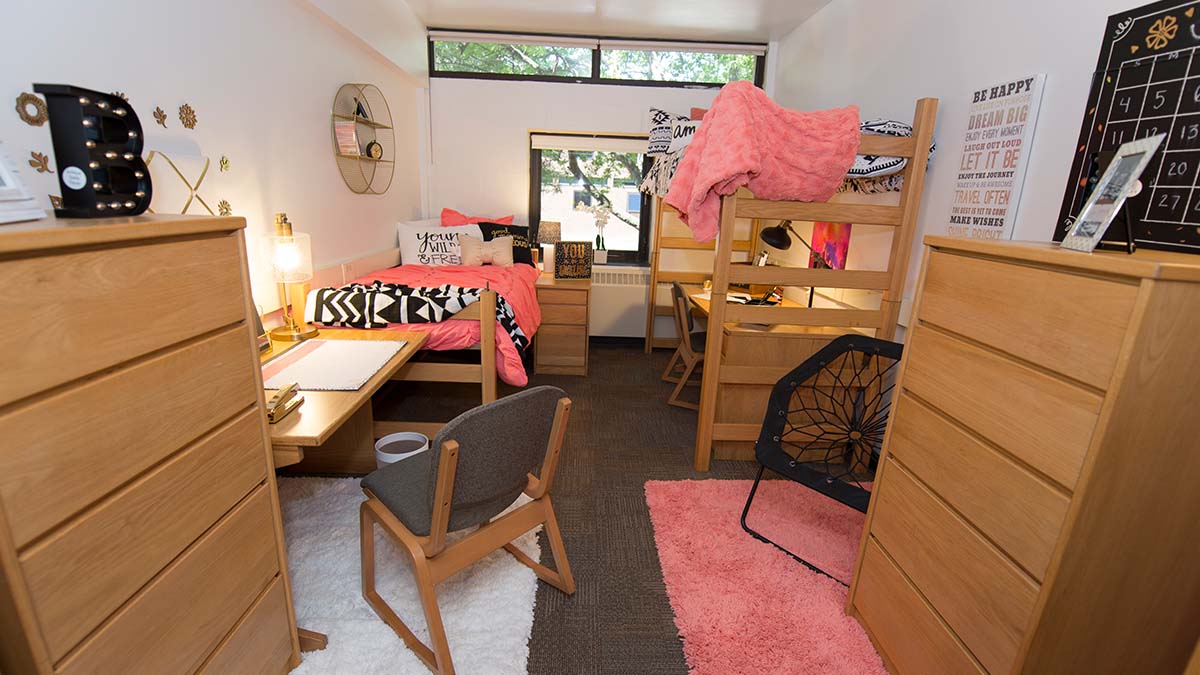 A dorm room with beds, dresses, desks, and pink decor.