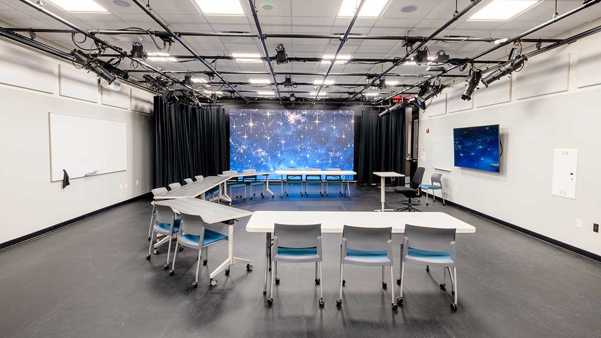 a large space with a lighting rig on the ceiling, screen, curtains, and U shaped table and chairs.