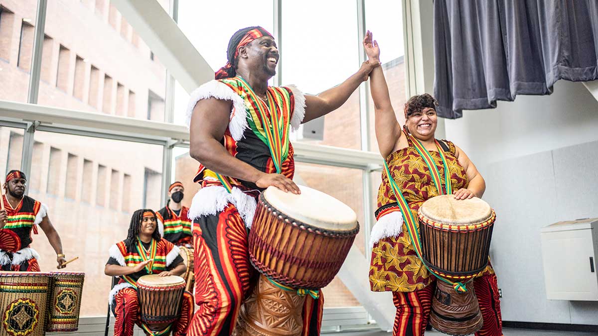 a group wearing traditional African clothing performs on tradition African drums, with a man and woman in the foreground, the man raising the woman's arm in celebration.