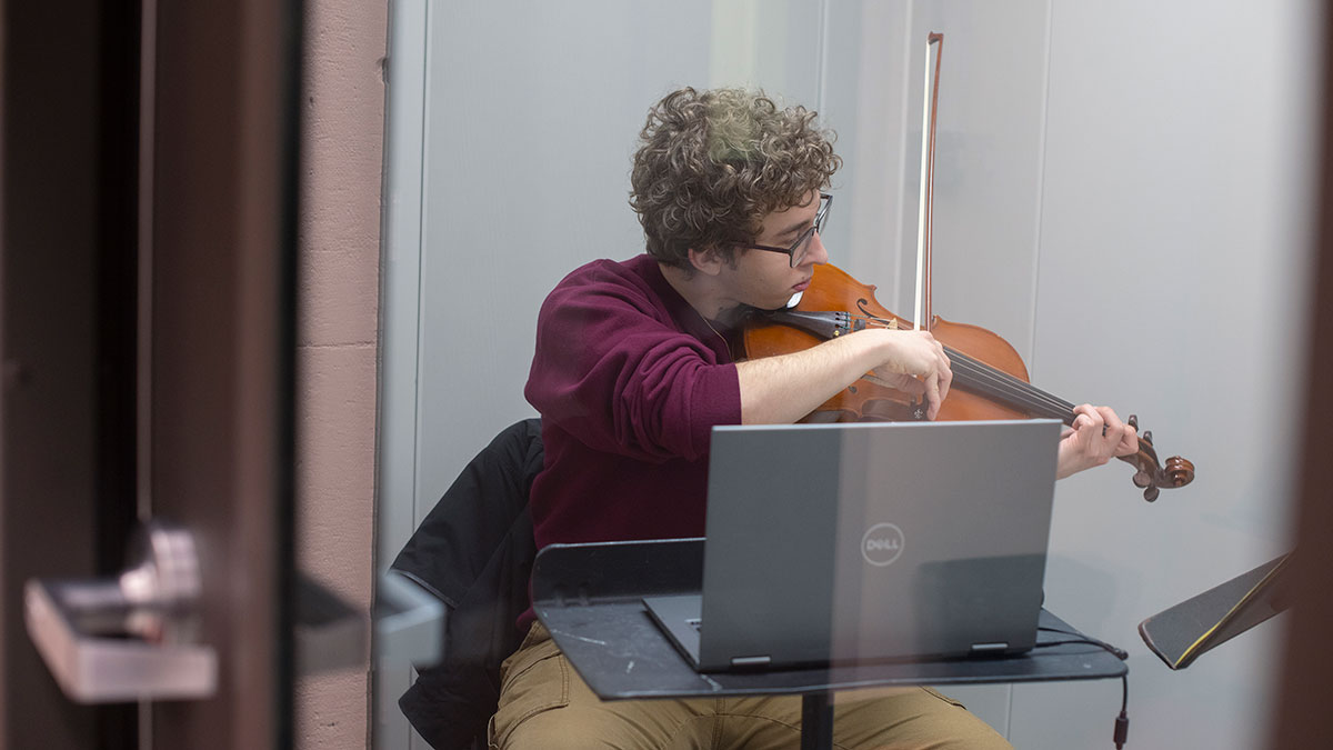 student playing the violin while looking at a laptop perched on a music stand.