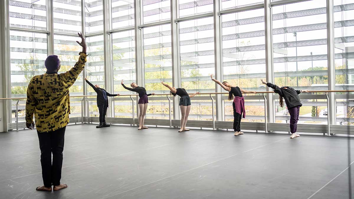 a line of five ballet dancers practicing on barre, with an instructor in front in a large, open space.