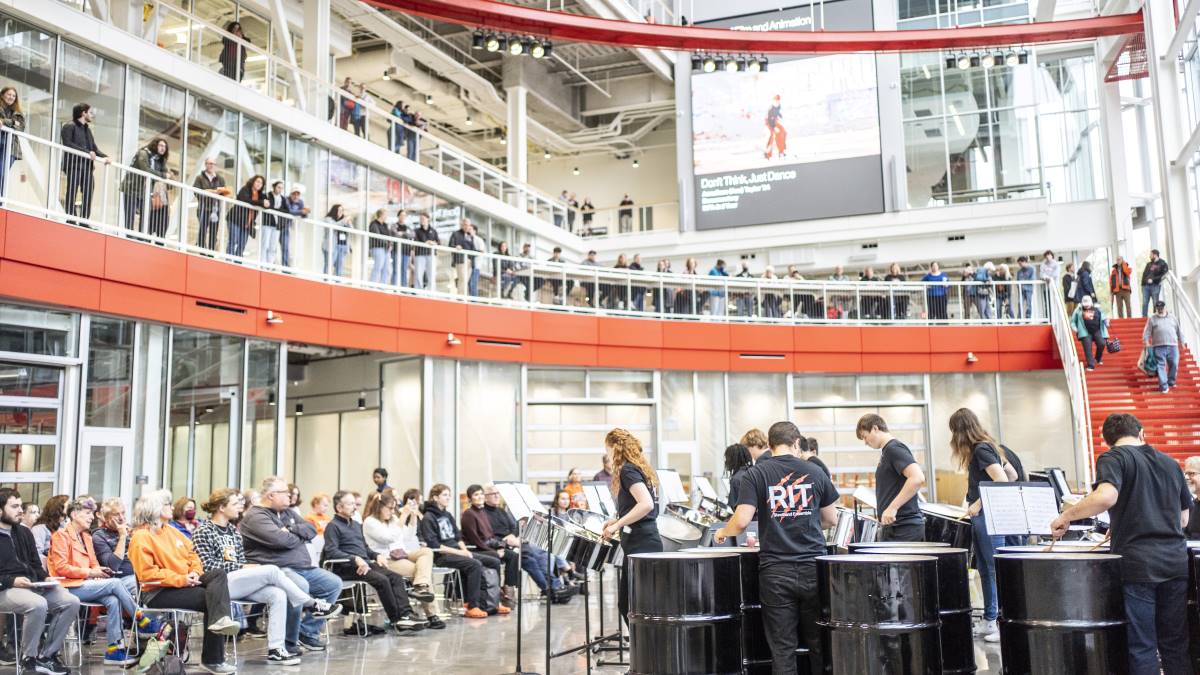 The R I T steel drum band performing in a large, open space, with audience members watcing from the ground floor and second floor.