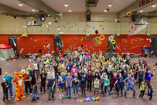 Large group of people throws juggling pins, hoops and balls into the air.