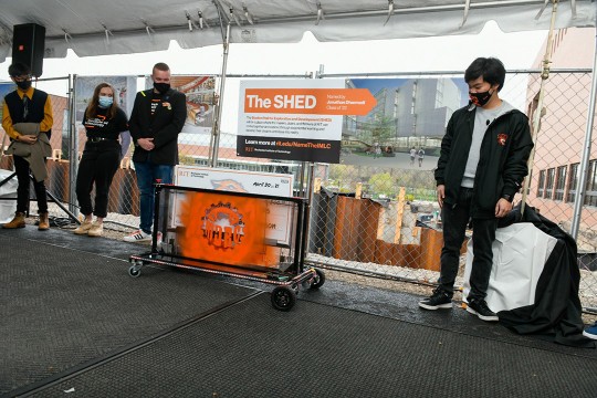 four students standing next to sign naming a building The Shed.