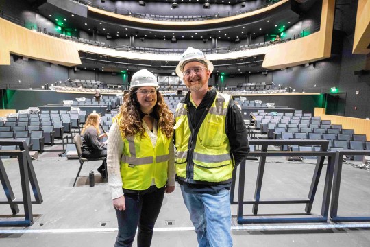 a woman and a man in construction gear smile for the camera in a performing arts auditorium.