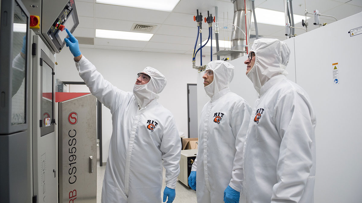 3 people in a clean room wearing white protective suits.