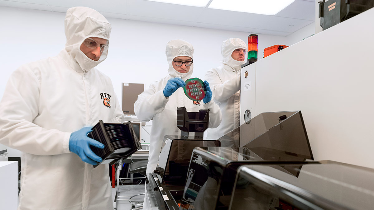three researchers in clean suits working with semiconductors.