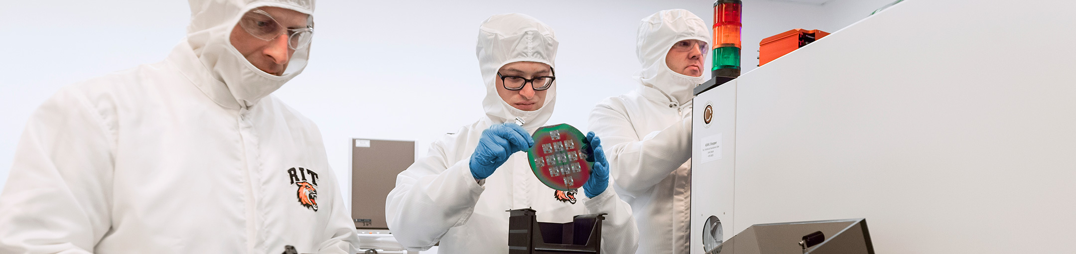 3 people in a clean room wearing white protective suits.