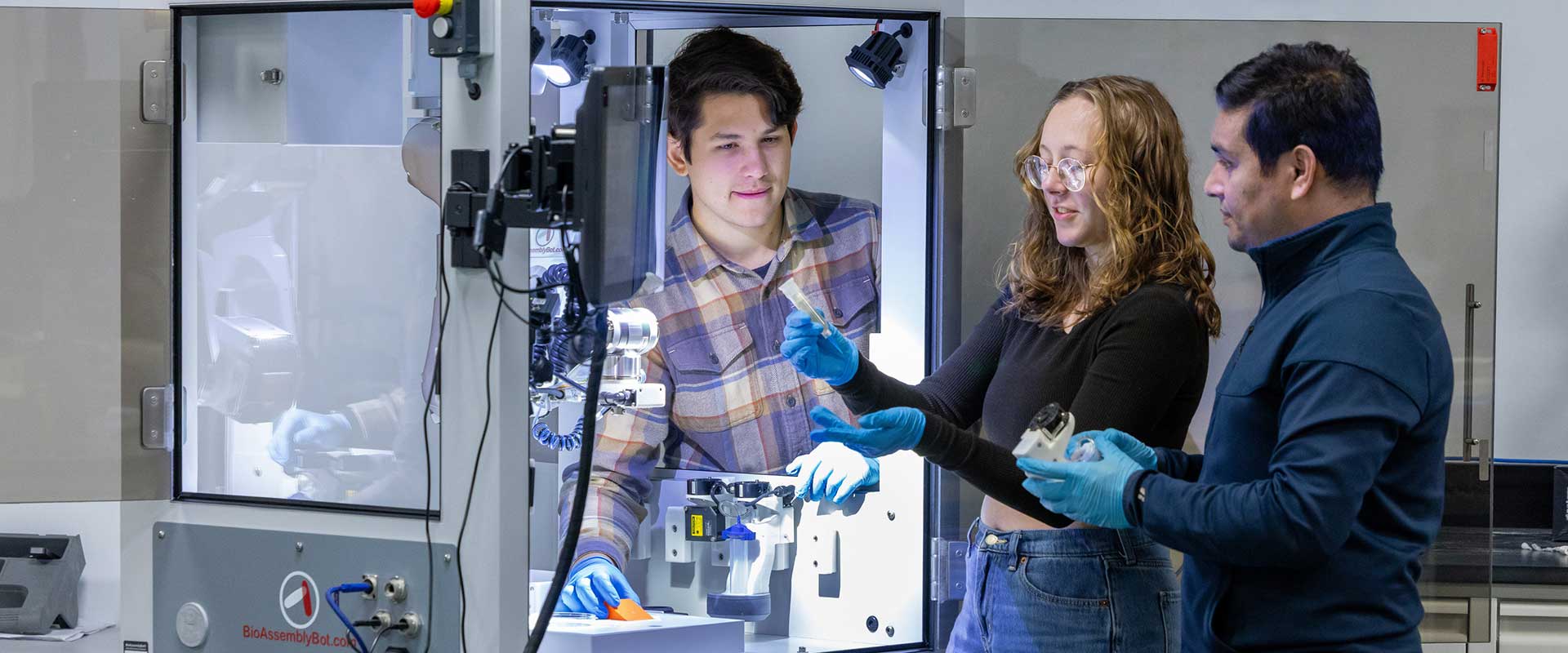 two students and a professor look at samples near a large 3 D printer device.