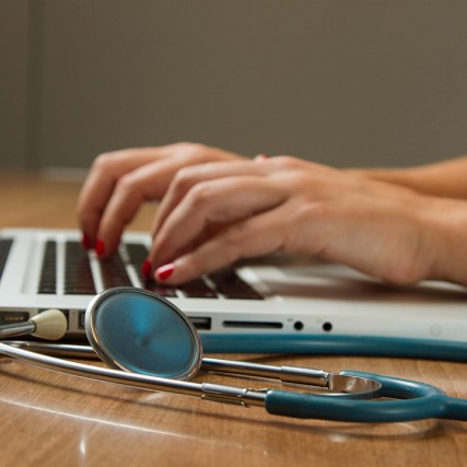 hands typing on a laptop next to a stethoscope.