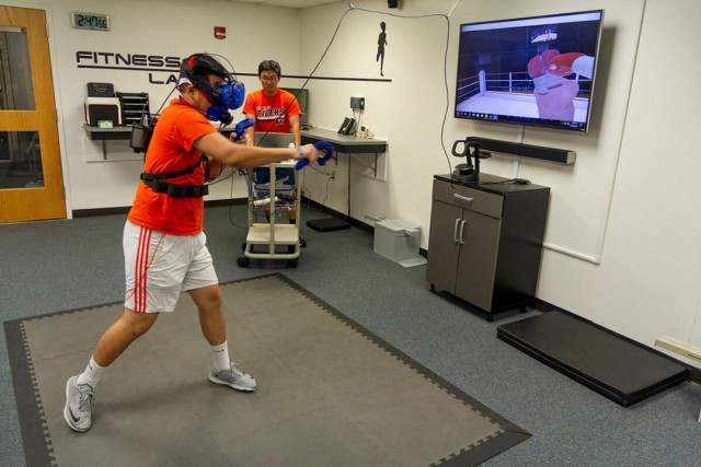 college student wearing a virtual reality headset and throwing a punch. A T V screen shows a simulation of a boxing ring.