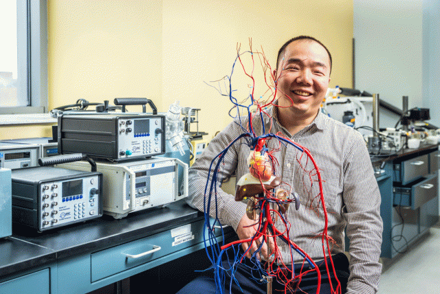 Smiling man in labratory holds a detailed anatomical model of a human heart with red and blue blood vessels. Electronic testing equipment and lab instruments are visible on the bench behind him.