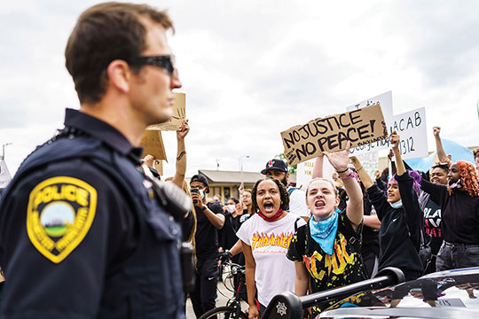 A police officer standing in front of a crowd holding a sign that says No Justice No Peace