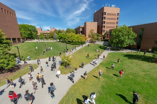 'Students walking across walkway and lawn between brick buildings.'