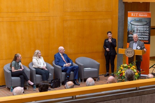 'Man speaks at podium with interpreter next to him and three people seated against a wall.'